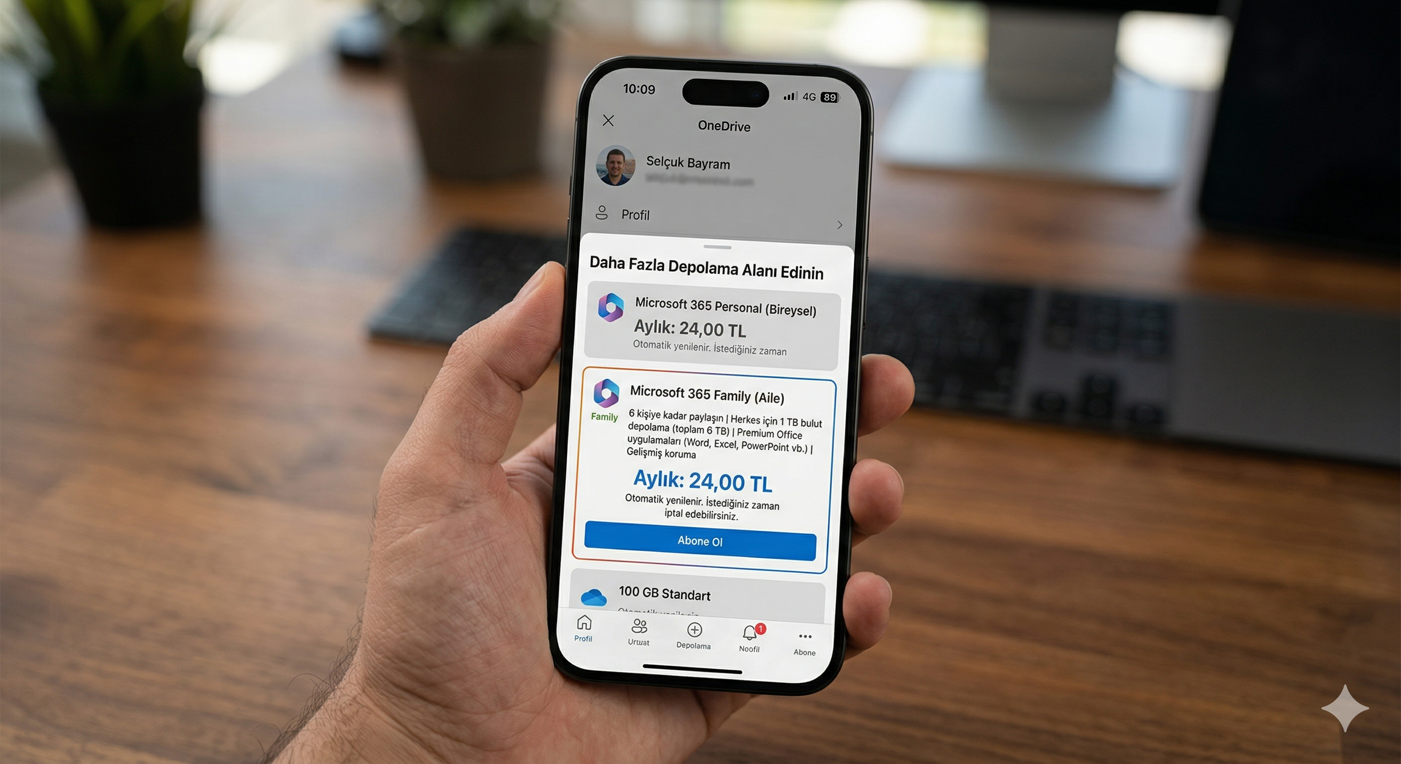 Net olmayan bir ofis masası önünde, bir kişinin elinde tuttuğu iPhone ekranı görülüyor. Telefondaki OneDrive uygulamasında 'Daha Fazla Depolama Alanı Edinin' başlıklı abonelik menüsü açık. Ekranın merkezinde yer alan 'Microsoft 365 Family (Aile)' seçeneği mavi bir çerçeveyle vurgulanmış; altında 'Aylık: 24,00 TL' ibaresi ve en altta mavi renkli belirgin bir 'Abone Ol' butonu yer alıyor."